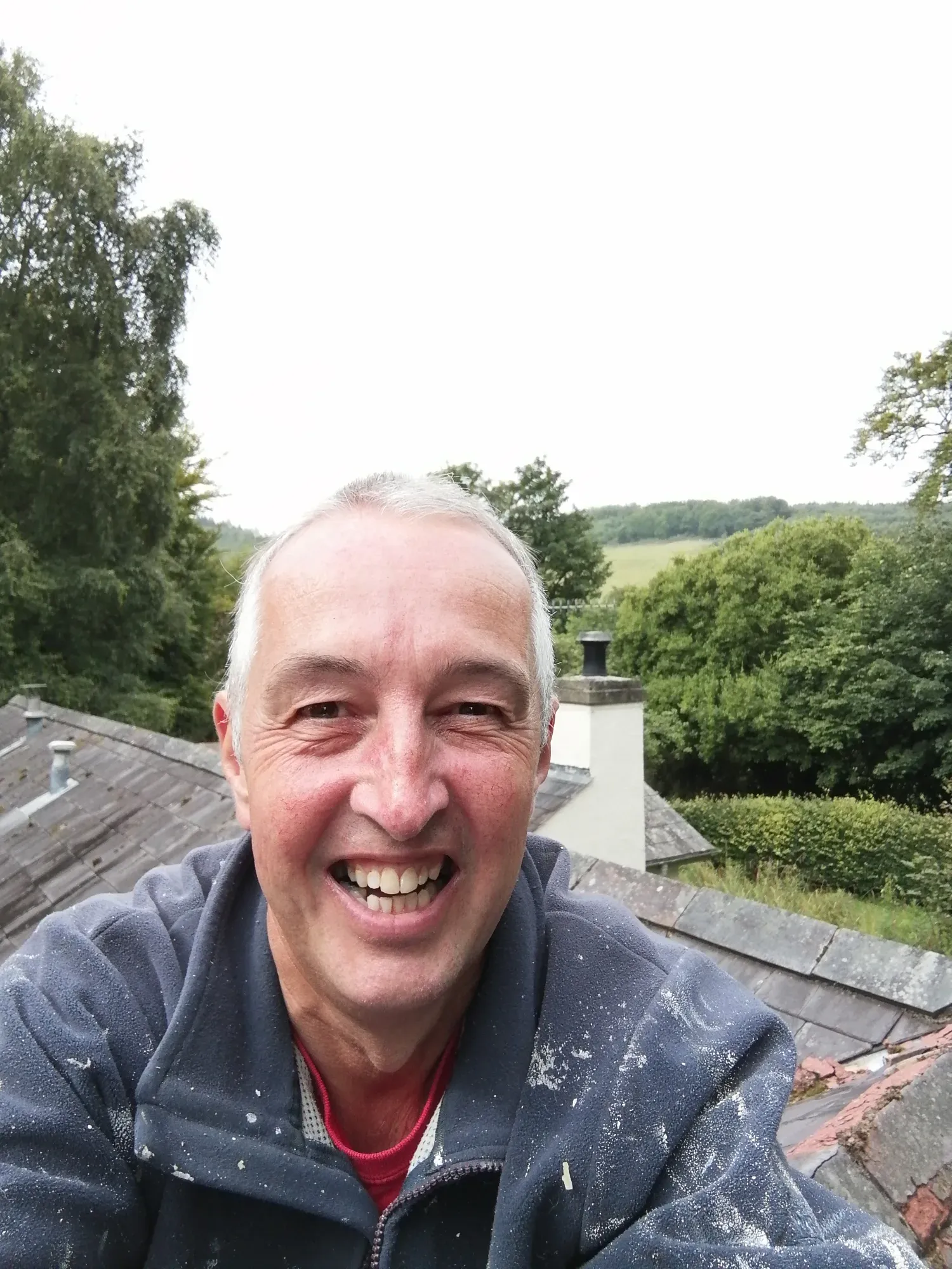 Ken Sives smiling on a roof, with a chimney and trees behind